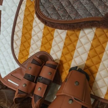 Um close-up do equipamento equestre Cloches Classic Sixteen Cypress, com uma almofada de sela acolchoada com listras douradas e brancas, botas de montaria de couro marrom e uma almofada de lã marrom com padrão de espinha de peixe.