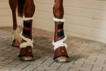 Close-up da parte inferior das pernas de um cavalo usando Cloches Sixteen Cypress com Carneira, botas protetoras marrons com forro de lã branca, parado em um piso pavimentado diante de uma parede de madeira branca.