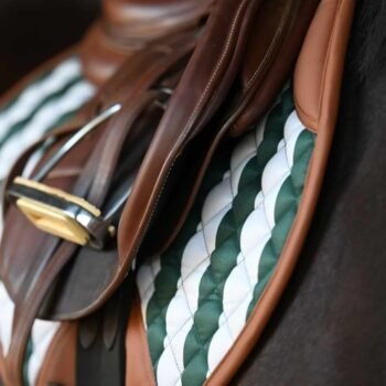 Close-up de uma sela de couro marrom em um cavalo com o protetor de sela Suadouro Classic Stripe Jump 16 Cypress, com um padrão listrado verde e branco, visível por baixo.