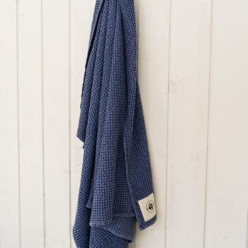 A Toalha de secagem e arrefecimento Waffle Sixteen Cypress azul, com uma pequena etiqueta branca, está pendurada em uma parede de madeira branca.